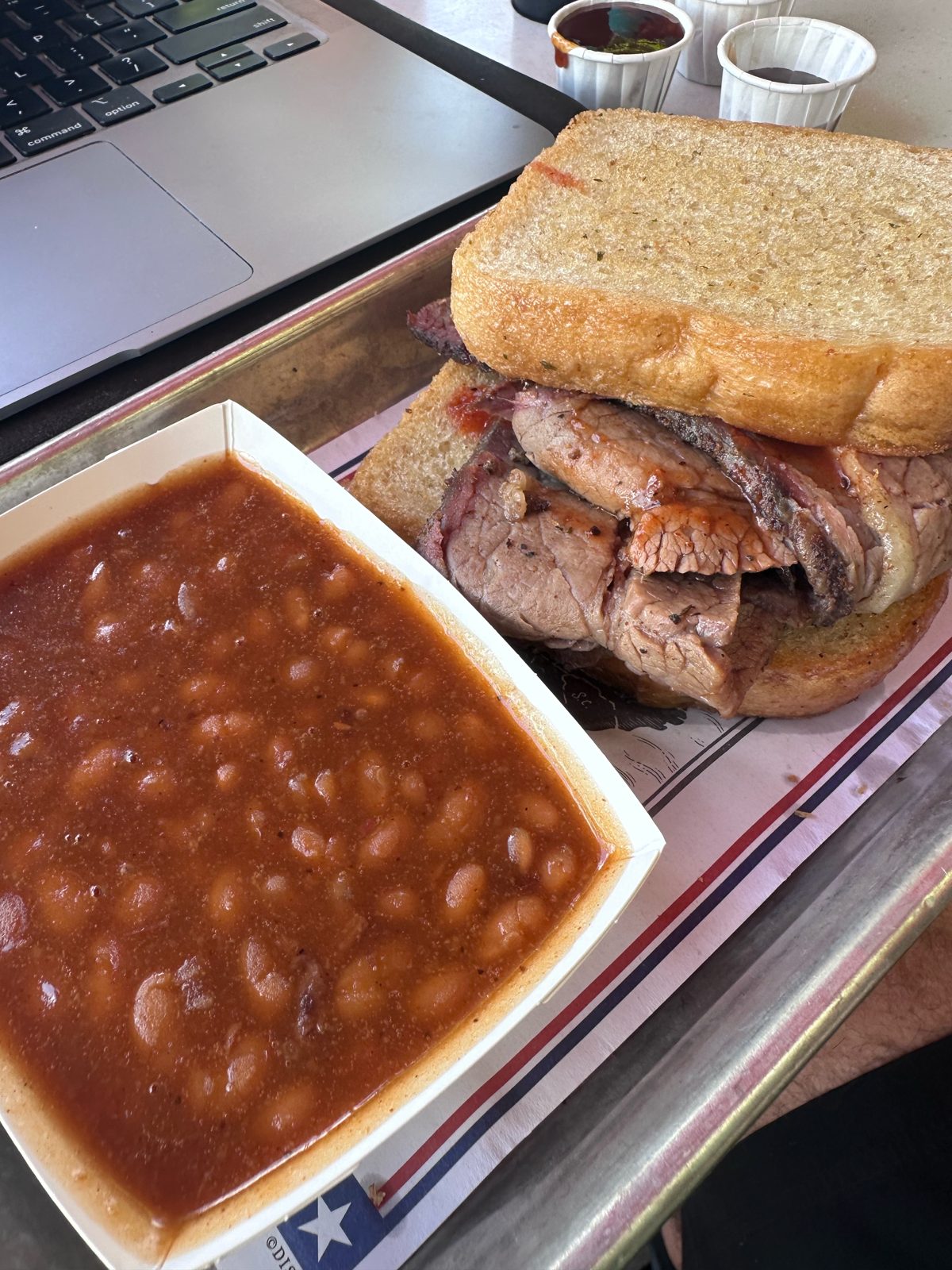 Texas brisket sandwich on Texas toast with a side of baked beans from Regal Eagle Smokehouse, on a tray next to a laptop