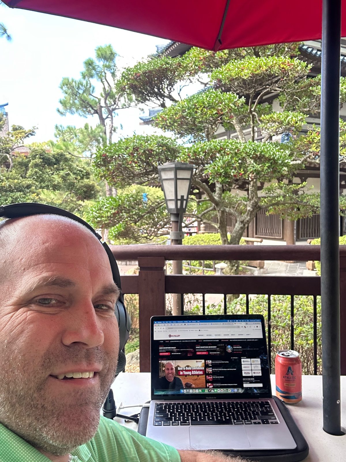 Ryan with his laptop under a red umbrella, pine trees and pagoda behind