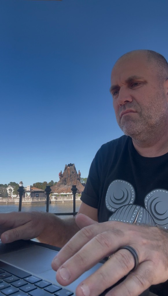 Ryan working on a laptop lakeside at Disney Springs with Rainforest Cafe across the water